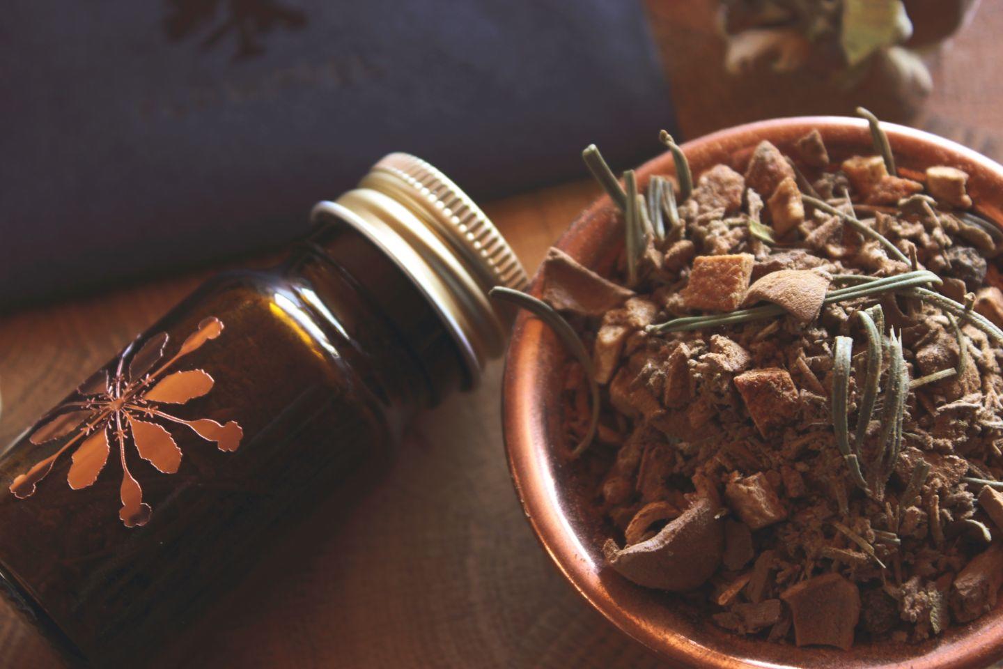 Kleine Braunglasflasche mit Hollenkraut-Logo, gefüllt mit Räucherwerk, liegend neben einer Schale mit getrockneten Kräutern und Pflanzenstücken auf einem warm beleuchteten Holzhintergrund.