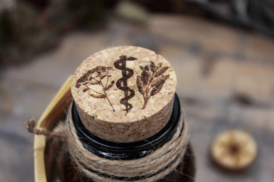 Korken in einer braunen Flasche. Auf dem Korken ist ein Motiv eingebrannt. Es zeigt in der Mitte den Asklepiosstab, links daneben Schafgarbe und rechts daneben Salbei.