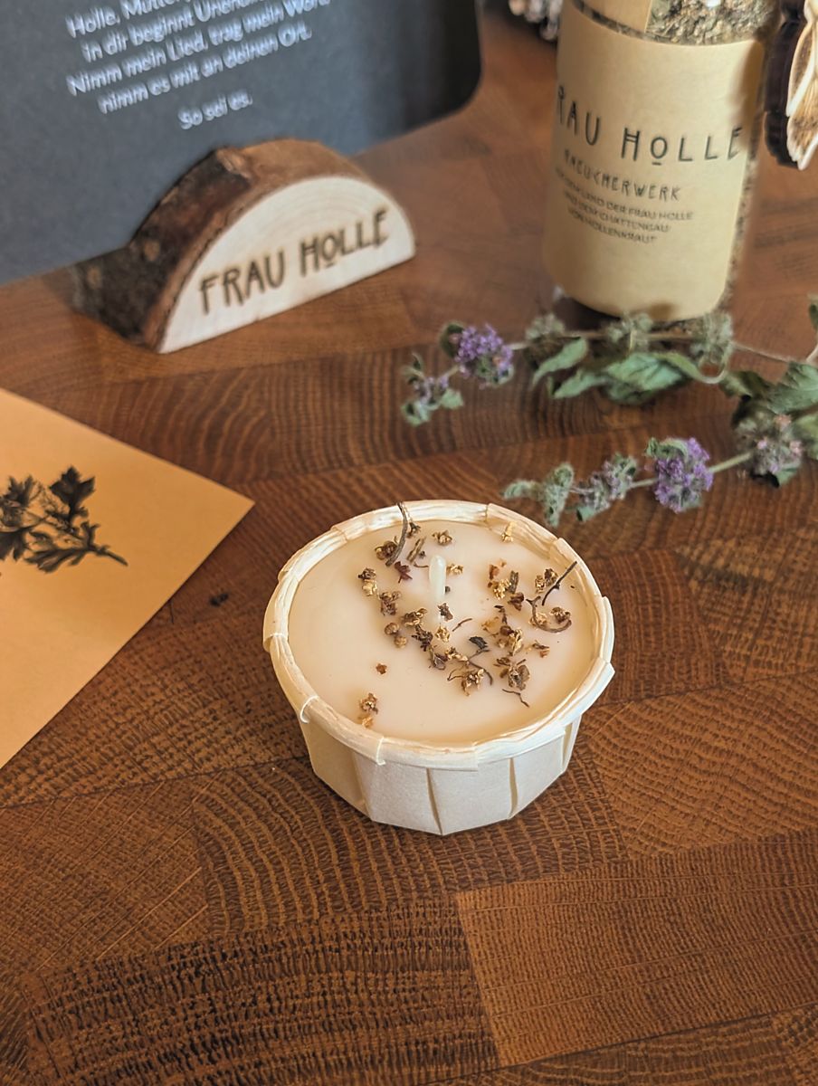 Die Ritualkerze mit Holunder aus Rapswachs. Sie hat in etwa die Größe eines Teelichts, ist aber in nicht brennbaren Papier eingeschlagen.