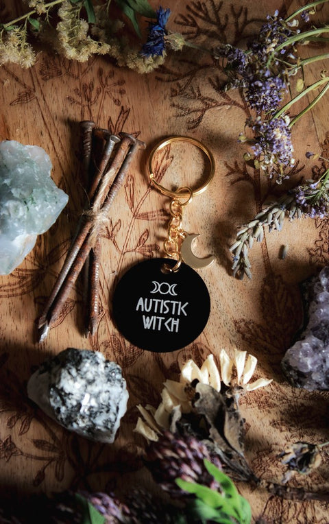 Ein Schlüsselanhänger in Schwarz und Gold mit goldenem Mond-Charm. Auf der schwarzen Scheibe steht in weiß: Autistic Witch