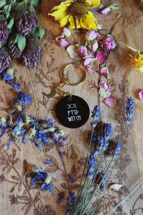 Das Bild zeigt eine Holzoberfläche mit floralen Gravuren, auf der getrocknete Blumen wie lila Klee, gelbe Sonnenblumen, rosa Rosenblätter und Lavendel arrangiert sind. Im Mittelpunkt liegt ein Schlüsselanhänger mit einem goldenen Ring und einem schwarzen, runden Anhänger. Auf dem Anhänger ist ein weißer Text mit der Aufschrift "PTSD WITCH" zu sehen, darüber befindet sich ein Triple-Mond-Symbol.