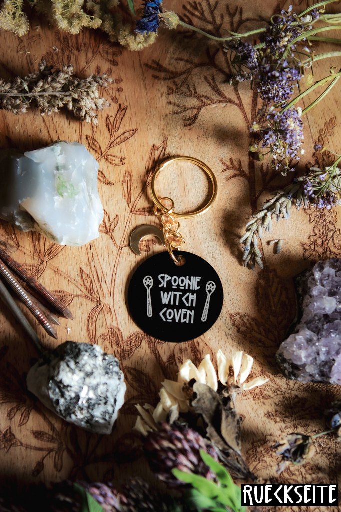 Ein Schlüsselanhänger in Schwarz und Gold mit goldenem Mond-Charm. Auf der schwarzen Scheibe steht in weiß: Spoonie Witch Coven.