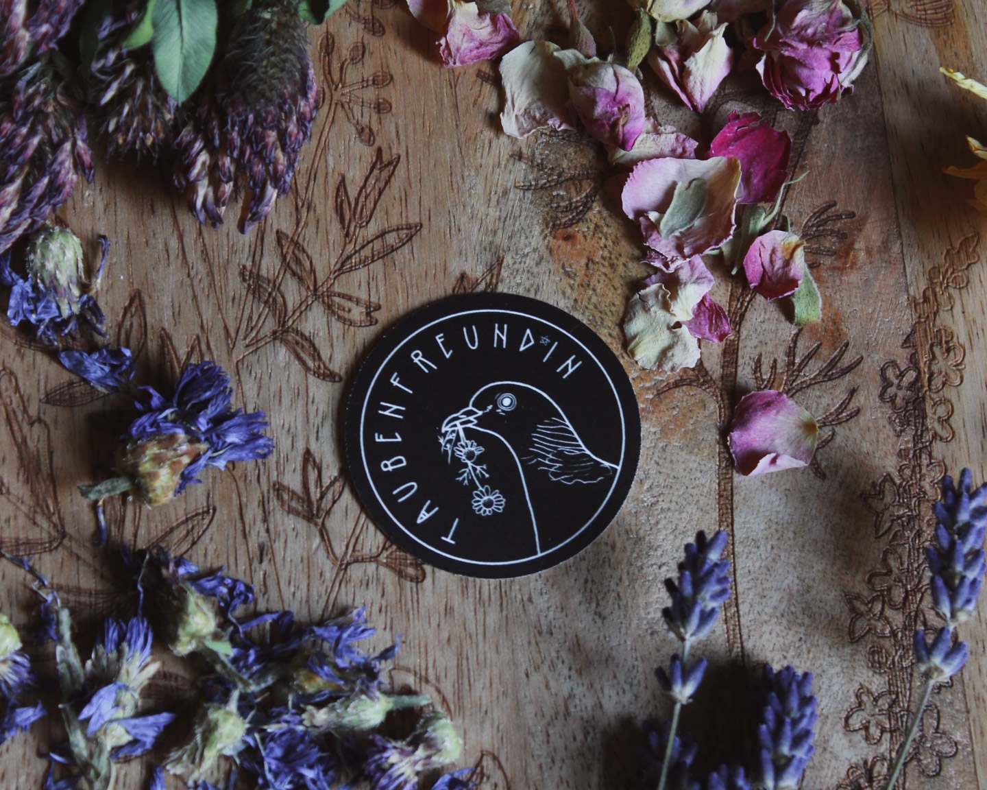 Das Bild zeigt eine Holzoberfläche mit kunstvollen floralen Gravuren. Darauf sind verschiedene getrocknete Blumen zu sehen, darunter lila und pinke Blütenblätter. In der Mitte des Arrangements befindet sich ein schwarzer, runder Sticker mit einer weißen Illustration, die eine Ringeltaube zeigt, die Kamille im Schnabel hält. Der Sticker trägt die Aufschrift „TAUBENFREUND*IN.“