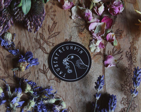Das Bild zeigt eine Holzoberfläche mit kunstvollen floralen Gravuren. Darauf sind verschiedene getrocknete Blumen zu sehen, darunter lila und pinke Blütenblätter. In der Mitte des Arrangements befindet sich ein schwarzer, runder Sticker mit einer weißen Illustration, die eine Ringeltaube zeigt, die Kamille im Schnabel hält. Der Sticker trägt die Aufschrift „TAUBENFREUND*IN.“