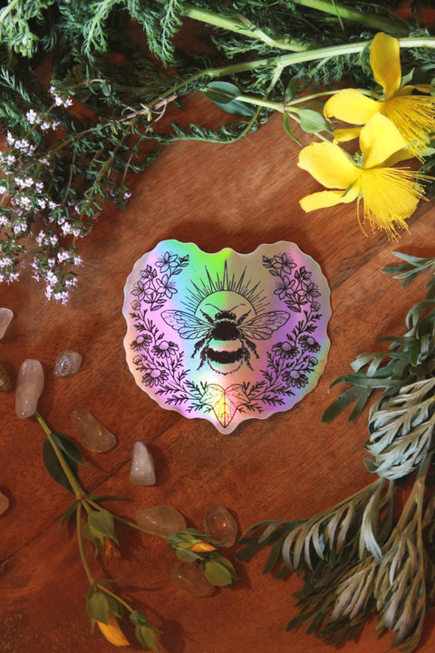 Auf einem Holztisch liegen verschiedene Kräuter wie Schafgarbe, Wermut und Johanniskraut um einen holographischen Aufkleber drumherum. Der Sticker zeigt eine Hummel. Eingerahmt wird sie von Johanniskraut, Kamille und einem Efeublatt.
