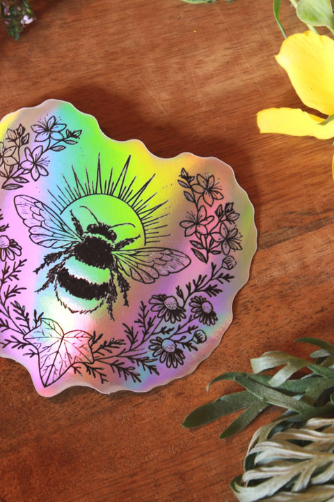 Nahaufnahme: Auf einem Holztisch liegen verschiedene Kräuter wie Schafgarbe, Wermut und Johanniskraut um einen holographischen Aufkleber drumherum. Der Sticker zeigt eine Hummel. Eingerahmt wird sie von Johanniskraut, Kamille und einem Efeublatt.