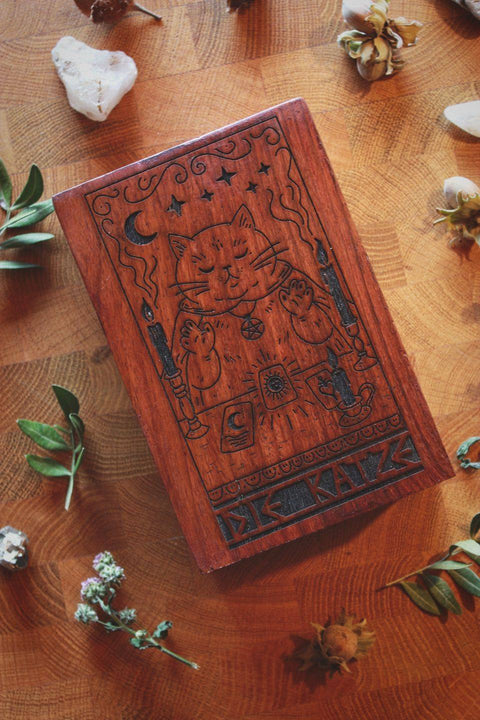 Holzkiste für Tarotkarten mit eingebranntem Motiv: Eine ruhende, mystisch wirkende Katze sitzt zwischen zwei brennenden Kerzen und legt Tarotkarten. Über ihr schweben Sterne und ein Mond, darunter steht ‚DIE KATZE‘. Die Szene ist von Blättern, Steinen und Naturmaterialien umgeben.