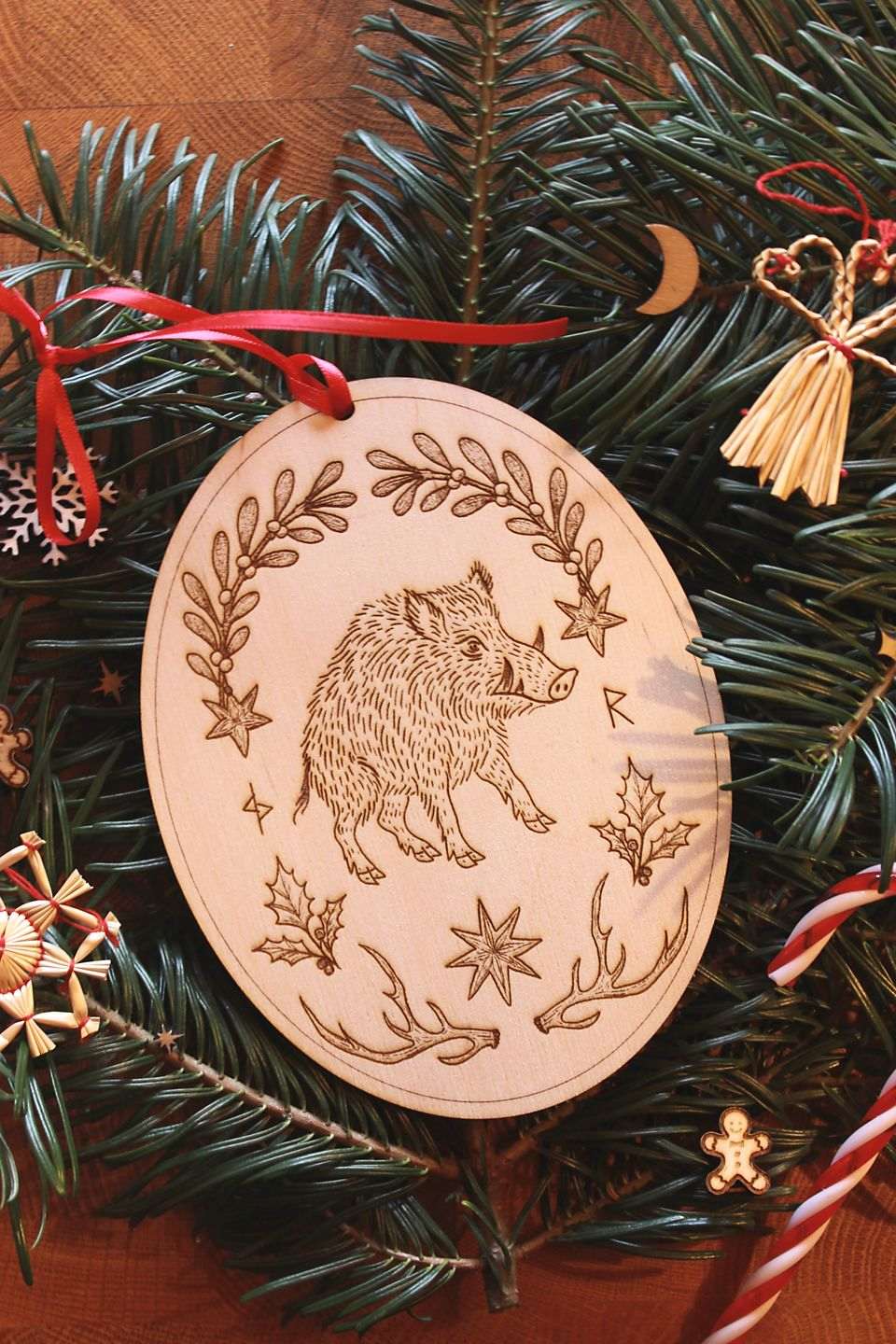 Hochkant: Gebettet auf Tannenzweigen und weihnachtliche Dekoration liegt in der Mitte ein ovales Ornament aus Holz. Diese Dekoration zu Jul (Wintersonnenwende) zeigt ein Wildschwein. Eingerahmt wird es von typischen Jul-Motiven wie Mistelzweigen, Stechpalmen, Strohsternen, Geweihen und passenden Runen.