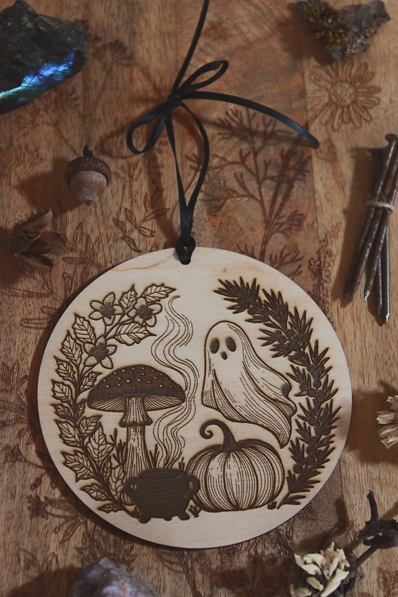 Hochkant: Rundes Holzornament mit filigranem Laserdesign: Neben Geist, Kürbis und Fliegenpilz steht ein Hexenkessel aus dem Rauch empor steigt. Eingerahmt ist das Motiv von schwarzem Bilsenkraut und Wacholder, die typisch für die Samhain-Nacht sind. Hängend an schwarzem Band, dekorativ umgeben von Naturmaterialien wie Steinen, getrockneten Blüten und Zweigen – ideal als saisonale Deko zu Samhain oder Halloween.