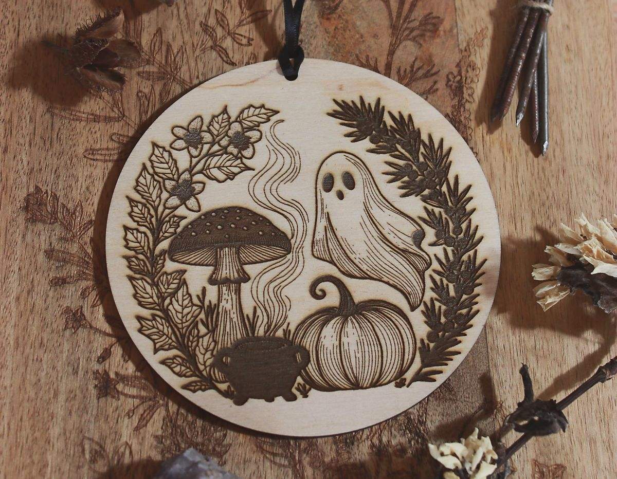 Rundes Holzornament mit filigranem Laserdesign: Neben Geist, Kürbis und Fliegenpilz steht ein Hexenkessel aus dem Rauch empor steigt. Eingerahmt ist das Motiv von schwarzem Bilsenkraut und Wacholder, die typisch für die Samhain-Nacht sind. Hängend an schwarzem Band, dekorativ umgeben von Naturmaterialien wie Steinen, getrockneten Blüten und Zweigen – ideal als saisonale Deko zu Samhain oder Halloween.