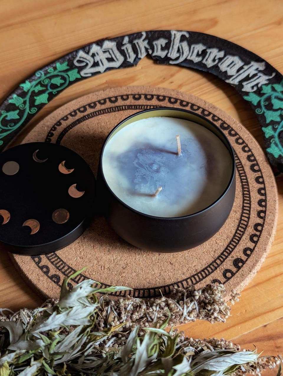 Das Bild zeigt ein rundes schwarzes Kerzengefäß, in dem sich eine blau-weiße Kerze mit zwei Dochten befindet, sowie ein schwarzer Deckel mit Symbolen der Mondphasen. Die Kerze steht auf einer dekorativen runden Korkplatte. Außerdem sind getrocknete Kräuter, eine geschwungene Sichel mit grünen Efeumotiven und das Wort „Witchcraft“ in gotischer Schrift zu sehen.