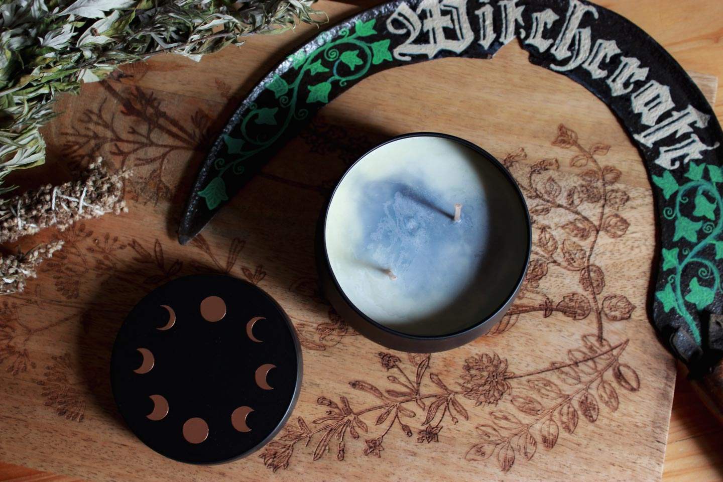 Das Bild zeigt eine Holzoberfläche mit botanischen Gravuren. Darauf befindet sich eine weiß-himmelblaue Kerze mit zwei Dochten sowie ein schwarzer Deckel mit Symbolen der Mondphasen. Außerdem sind getrocknete Kräuter, eine geschwungene Sichel mit grünen Efeumotiven und das Wort „Witchcraft“ in gotischer Schrift zu sehen.