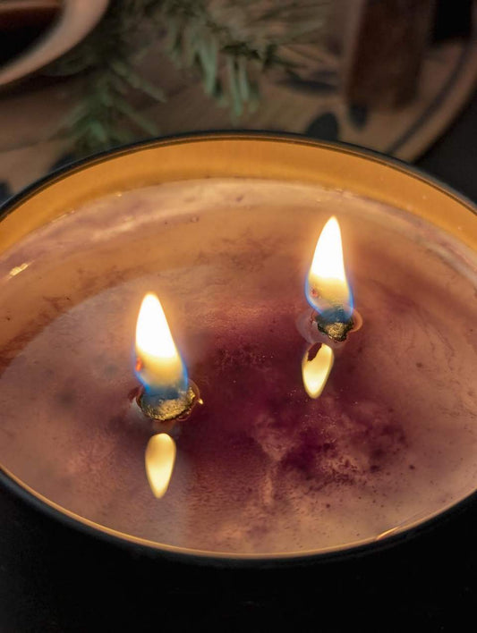 Das Bild zeigt eine Nahaufnahme einer angezündeten Kerze mit zwei Flammen. Die Kerze befindet sich in einem runden Behälter mit einem goldenen Rand. Das Wachs der Kerze hat einen rot-violetten Farbton, und die Flammen brennen hell mit einer blauen Basis und gelben Spitzen. Im Hintergrund sind einige grüne Kiefernadeln zu sehen.