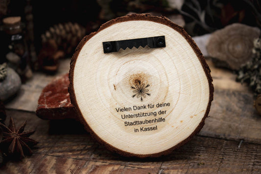 Rückseite Holzgravur-Taube-Taubenhilfe, Hollenkraut Logo mit Text "Vielen Dank für deine Unterstützung der Stadttaubenhilfe in Kassel, Aufhängung wurde befestigt, präsentiert in einer rustikalen Umgebung mit Kiefernzapfen,