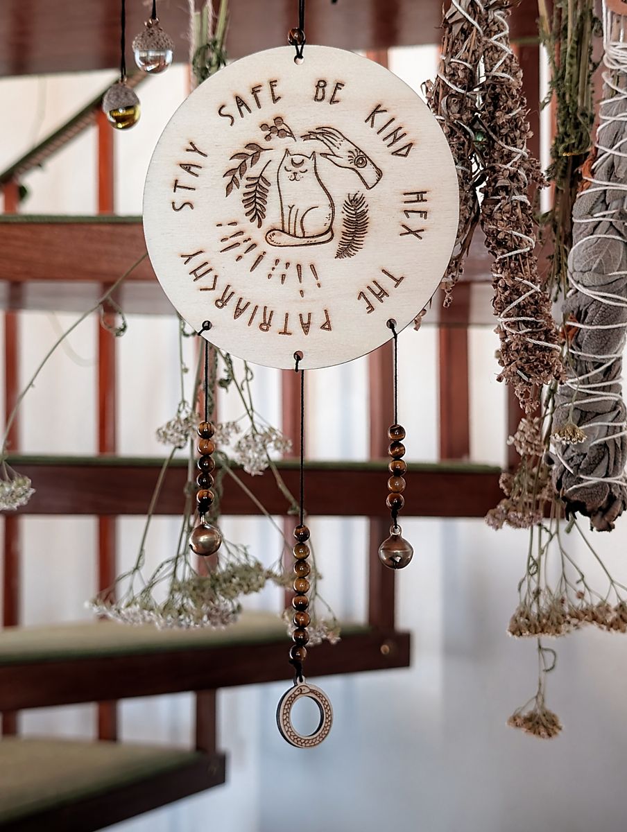 An einem Haken hängt eine Holzscheibe auf der ein Motiv eingebrannt ist. Es zeigt eine Katze, eine Hand und einige Pflanzen. Drumherum steht der Spruch "Stay safe be kind hex the patriarchy" An der Holzscheibe baumeln Glöckchen, eine Holzscheibe mit dem Motiv der Weltenschlange und Tigerauge.