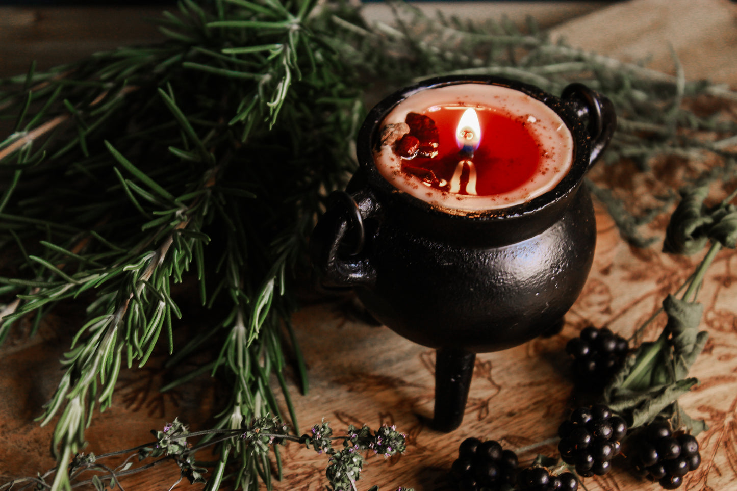 Schwarzer Kessel mit rotem Wachs, die Kerze wurde entzündet umgeben von Kräutern und Beeren, alles wurde schön auf einem braunen Brettchen präsentiert