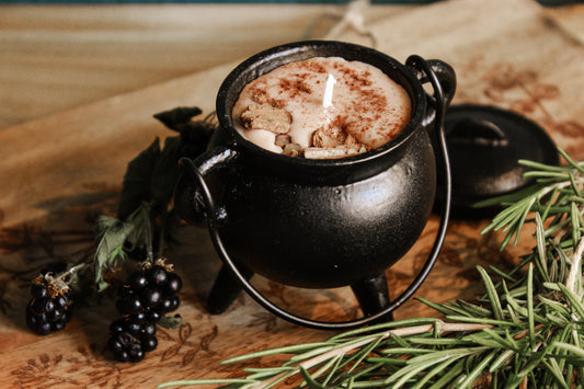 Schwarzer Kessel mit weißem Wachs, das Wachs ist leicht bräunlich gefärbt, umgeben von Kräutern und Beeren, alles wurde schön auf einem braunen Brettchen präsentiert