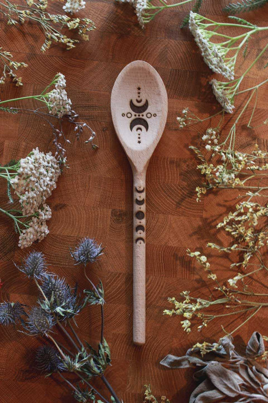 Hochkant: Von getrockneten Kräutern umgeben, liegt ein Kochlöffel aus Holz auf einem ebenso hölzernen Untergrund. Auf den Holzlöffel sind die Mondphasen eingebrannt.