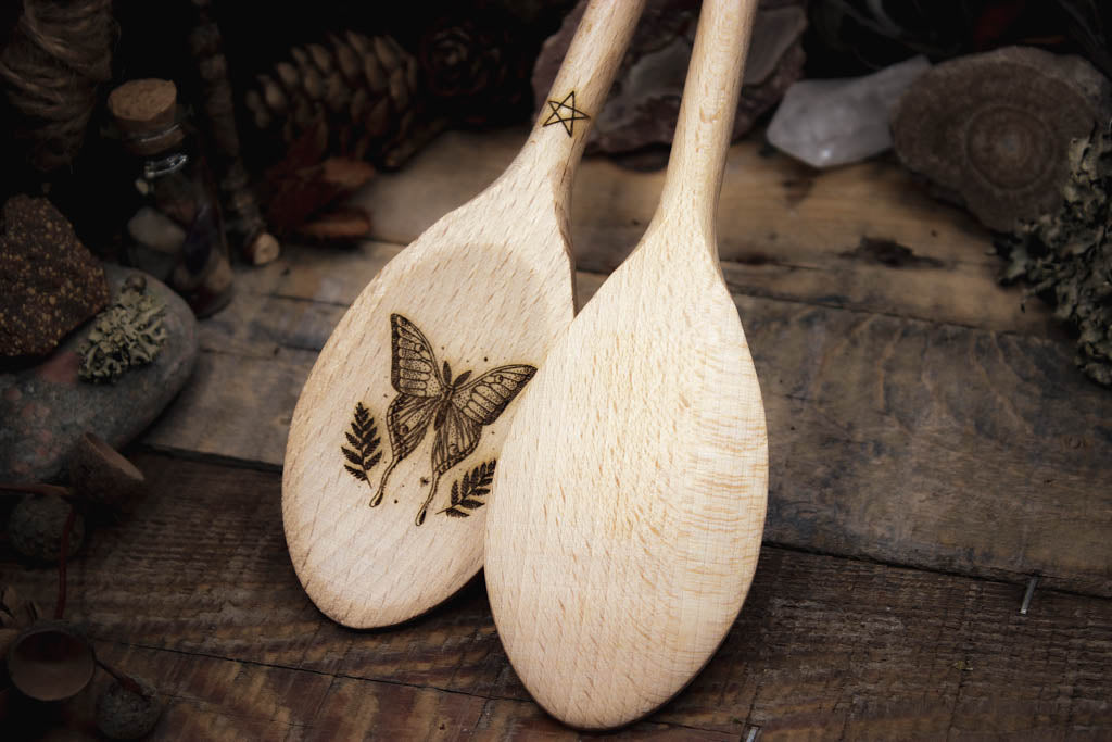 Zwei gravierte Holzlöffel mit kunstvoller Schmetterlings- und Blattgravur, präsentiert auf einem rustikalen Hintergrund mit getrockneten Blumen und Kiefernzapfen.