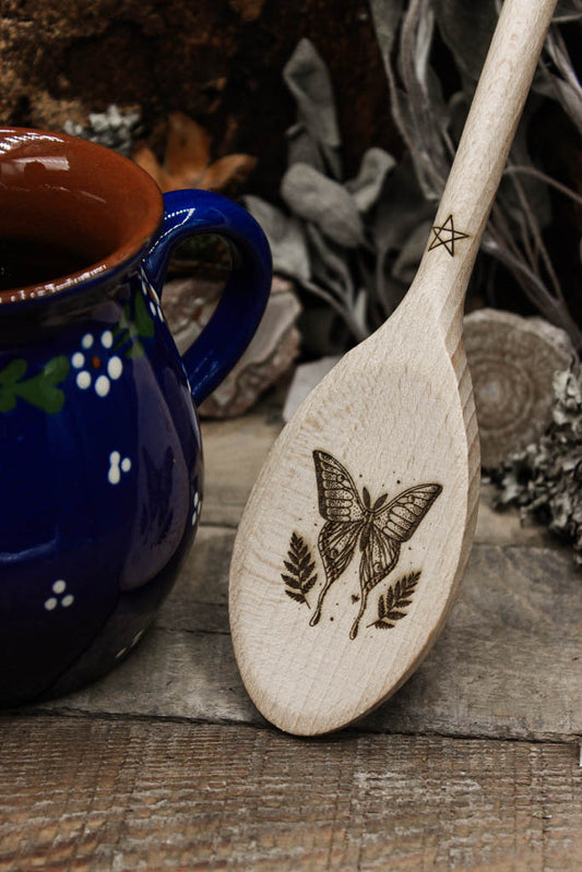 Handgeschnitzter Holzlöffel mit kunstvoller Schmetterlings- und Blattgravur, präsentiert auf einem rustikalen Hintergrund mit getrockneten Blumen und Kiefernzapfen. man sieht eine blaue Kanne als Dekoration daneben stehen