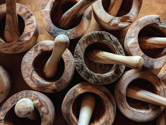 Foto von oben: Mehrere Mörser & Stößel aus Olivenholz stehen auf einem hölzernen Untergrund. Sie sehen rustikal aus und haben teilweise noch Rinde an sich.