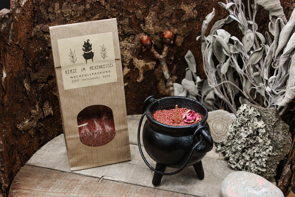 Eine Tüte Nachfüllpackung für die Kerze im Hexenkessel mit rotem Wachs darin. Im Vordergrund steht ein Kessel mit rotem Wachs.