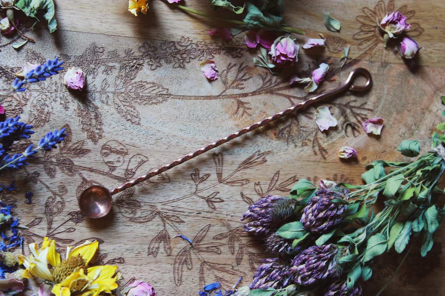 Das Bild zeigt eine rustikale, künstlerische Komposition auf einer Holzoberfläche mit filigranen floralen Gravuren. Darauf arrangiert sind verschiedene getrocknete Blumen und Kräuter wie Lavendel, Rosen und andere bunte Blüten. In der Mitte des Bildes liegt ein langes, gedrehtes Metalllöffelchen aus Kupfer mit einer kleinen, runden Schale an einem Ende. Die Kombination der Blumen, Kräuter und des Löffelchens auf der kunstvollen Holzoberfläche schafft eine harmonische, ästhetische Stimmung.