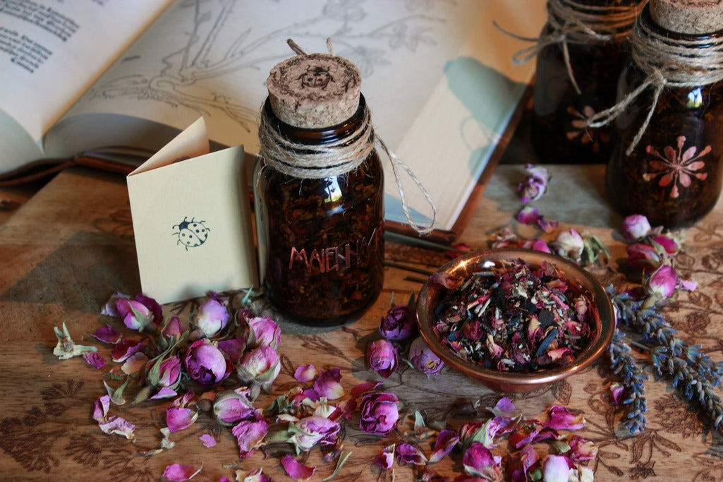 Drei Braunflaschen mit Korken, der mit einem Marienkäfer versehen ist, stehen auf einem Holztisch. Drumherum sind Lavendel- und Rosenblüten. Das kupferfarbene Etikett ist der Schriftzug des Räucherwerks: Maiennacht. Neben den Gläsern steht eine kleine Schale aus Kupfer in der die Räuchermischung zu sehen ist. Im Hintergrund liegt ein aufgeschlagenes altes Kräuterbuch. Neben einem der Gläser steht ein Beizettel mit dem Stempel eines Marienkäfers darauf.