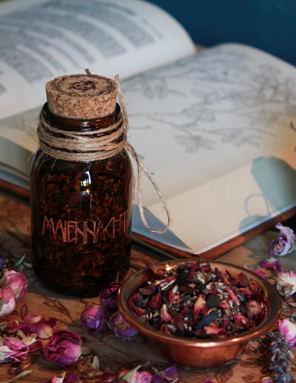 Eine Braunglas-Flasche mit Korken, der mit einem Marienkäfer versehen ist, steht auf einem Holztisch. Drumherum sind Lavendel- und Rosenblüten. Das kupferfarbene Etikett zeigt das Hollenkraut-Logo. Neben dem Glas steht eine kleine Schale aus Kupfer in der die Räuchermischung zu sehen ist. Im Hintergrund ist ein aufgeschlagenes altes Kräuterbuch zu sehen.