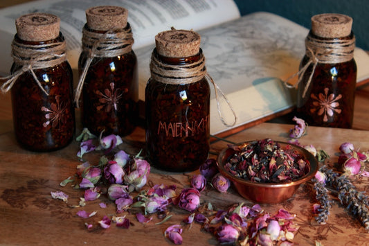 Vier Braunflaschen mit Korken, der mit einem Marienkäfer versehen ist, stehen auf einem Holztisch. Drumherum sind Lavendel- und Rosenblüten. Das kupferfarbene Etikett ist der Schriftzug des Räucherwerks: Maiennacht. Neben den Gläsern steht eine kleine Schale aus Kupfer in der die Räuchermischung zu sehen ist.