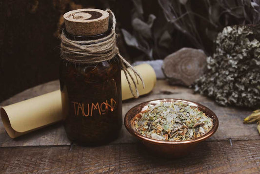 Auf einem Holzbrett steht eine braune Flasche mit Korken auf der in kupferfarbener Schrift Taumond steht. Oben auf dem Korken ist eine schwarze Mondsichel und eine Lindenblüte eingraviert. Hinter dem Glas liegt ein eingerollter Zettel. Daneben ist eine kupferfarbene Schale mit hellem Räucherwerk aus Kräutern, Blüten und einem weißem Harz zu sehen.