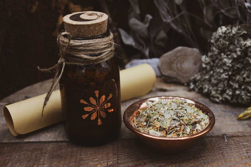 Auf einem Holzbrett steht eine braune Flasche mit Korken auf der das kupferfarbene Hollenkraut-Logo zu sehen ist. Oben auf dem Korken ist eine schwarze Mondsichel und eine Lindenblüte eingraviert. Hinter dem Glas liegt ein eingerollter Zettel. Daneben ist eine kupferfarbene Schale mit hellem Räucherwerk aus Kräutern, Blüten und einem weißem Harz zu sehen.
