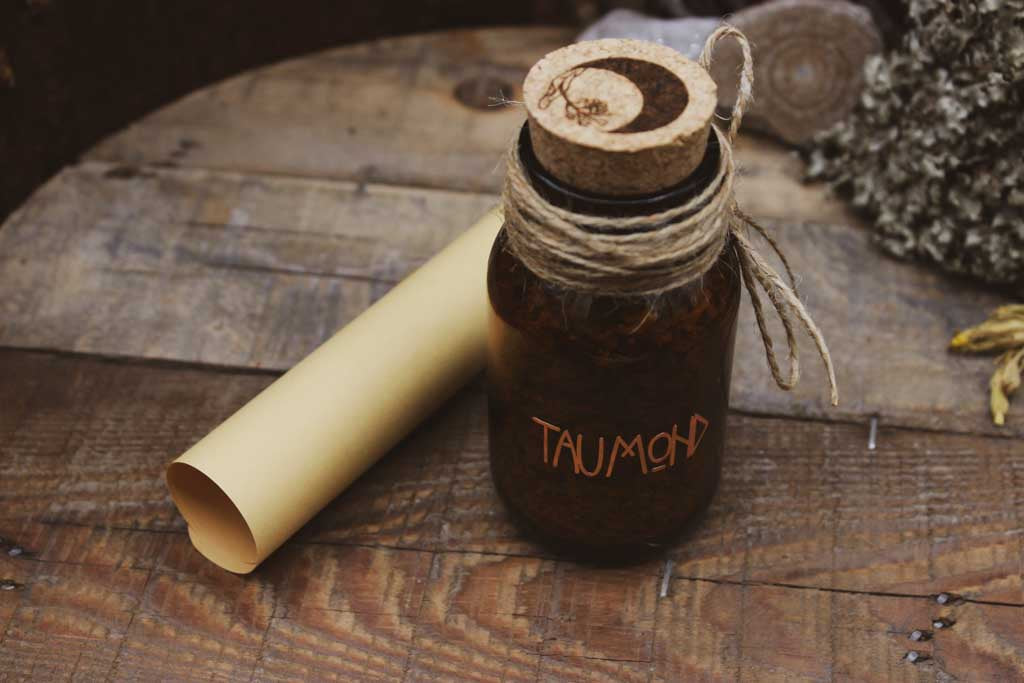 Auf einem Holzbrett steht eine braune Flasche mit Korken auf der in kupferfarbener Schrift Taumond steht. Oben auf dem Korken ist eine schwarze Mondsichel und eine Lindenblüte eingraviert. Hinter dem Glas liegt ein eingerollter Zettel.