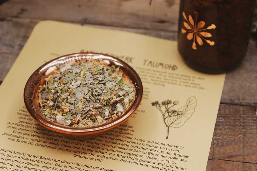 Ein beiges Blatt Papier liegt auf einem Holzbrett. Darauf zu sehen ist ein Text der das Räucherwerk Taumond beschreibt, sowie eine Zeichnung einer Lindenblüte. Auf dem Blatt steht eine kupferfarbene Schalle mit der Räucherung aus Kräutern und Harzen zu Imbolc.