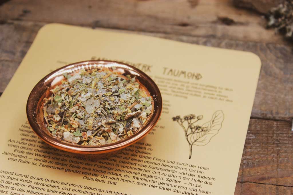 Ein beiges Blatt Papier liegt auf einem Holzbrett. Darauf zu sehen ist ein Text der das Räucherwerk Taumond beschreibt, sowie eine Zeichnung einer Lindenblüte. Auf dem Blatt steht eine kupferfarbene Schalle mit der Räucherung aus Kräutern und Harzen zu Imbolc.