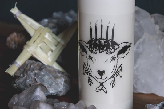 Nahaufnahme: Umringt von verschiedenen Mineralien und Steinen steht eine große champagner-farbene Stumpenkerze auf einem Holztisch. Darauf zu sehen ist ein schwarzes Motiv, das einen Kopf eines Schafes zeigt. Das Schaf trägt fünf schwarze Kerzen auf dem Haupt, umringt von mehreren Schlüsselblumenblüten. Unter dem Kopf sind fünf Schneeglöckchenblüten. Links hinter der Kerze steht ein Kreuz der Brigid aus Stroh.