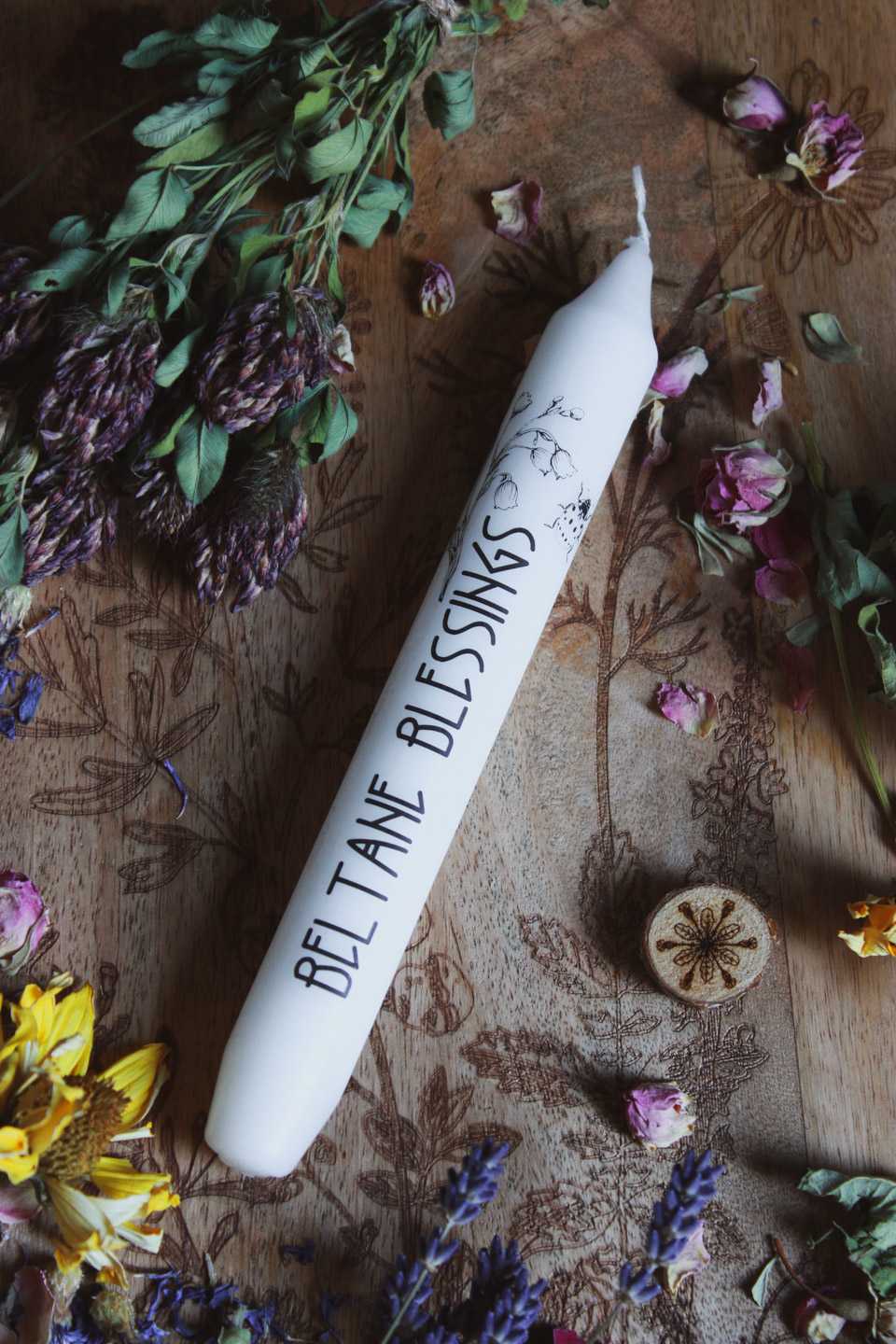 Hochkant: Das Bild zeigt eine weiße Kerze mit der Aufschrift „BELTANE BLESSINGS“ und dem Motiv von Maiglöckchen und einem Marienkäfer. Die Kerze liegt auf einer Holzfläche, die mit floralen Gravuren verziert ist. Rund um die Kerze sind getrocknete Blumen arrangiert, darunter gelbe, violette und pinke Blüten, sowie grüne Blätter.