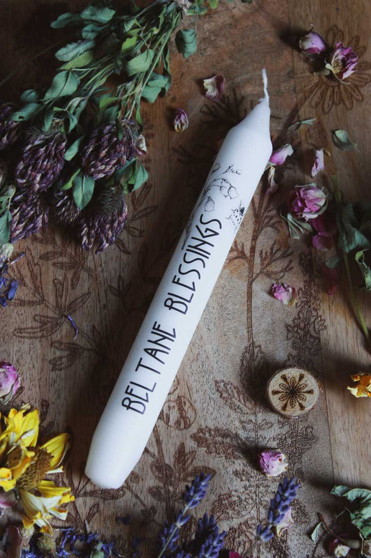 Hochkant: Das Bild zeigt eine weiße Kerze mit der Aufschrift „BELTANE BLESSINGS“ und dem Motiv von Maiglöckchen und einem Marienkäfer. Die Kerze liegt auf einer Holzfläche, die mit floralen Gravuren verziert ist. Rund um die Kerze sind getrocknete Blumen arrangiert, darunter gelbe, violette und pinke Blüten, sowie grüne Blätter.