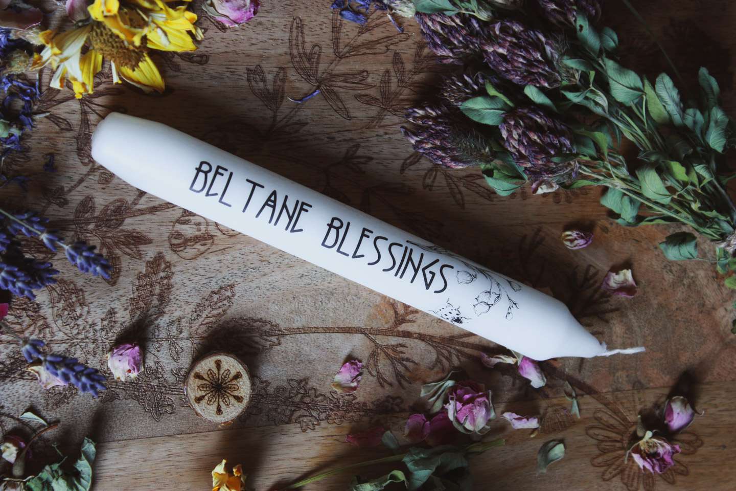 Das Bild zeigt eine weiße Kerze mit der Aufschrift „BELTANE BLESSINGS“ und dem Motiv von Maiglöckchen und einem Marienkäfer. Die Kerze liegt auf einer Holzfläche, die mit floralen Gravuren verziert ist. Rund um die Kerze sind getrocknete Blumen arrangiert, darunter gelbe, violette und pinke Blüten, sowie grüne Blätter.