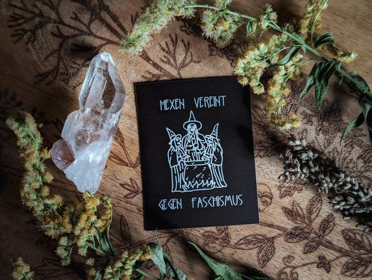 Von Pflanzen und einem Bergkristall umringt, liegt ein rechteckiger schwarzer Sticker mit weißem Aufdruck. Darauf zu sehen sind drei gezeichnete Hexen um einen Hexenkessel herum. Dazu der Text: Hexen vereint gegen Faschismus