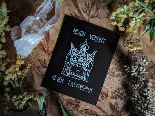Von Pflanzen und einem Bergkristall umringt, liegt ein rechteckiger schwarzer Sticker mit weißem Aufdruck. Darauf zu sehen sind drei gezeichnete Hexen um einen Hexenkessel herum. Dazu der Text: Hexen vereint gegen Faschismus