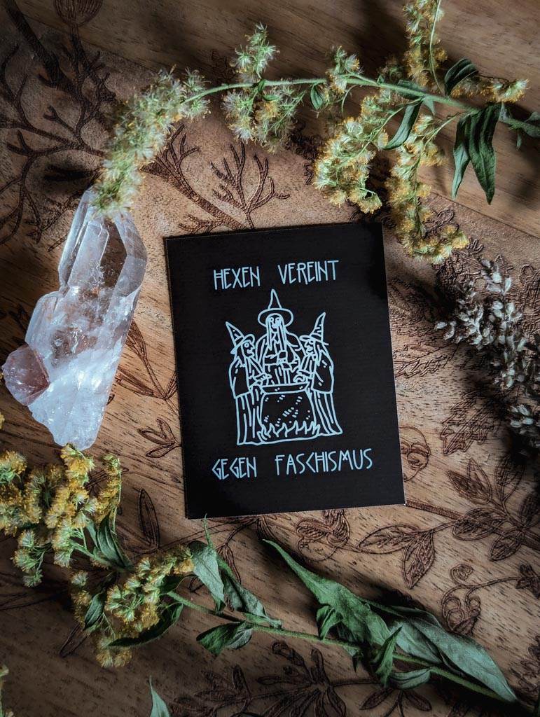 Von Pflanzen und einem Bergkristall umringt, liegt ein rechteckiger schwarzer Sticker mit weißem Aufdruck. Darauf zu sehen sind drei gezeichnete Hexen um einen Hexenkessel herum. Dazu der Text: Hexen vereint gegen Faschismus
