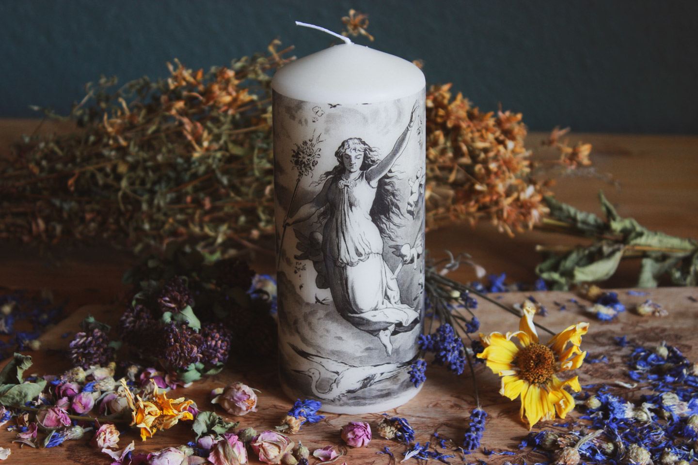 Das Bild zeigt eine fast weiße zylindrische Kerze mit einer detaillierten schwarz-weißen Illustration. Die Illustration zeigt eine Frau in einem fließenden Kleid, die einen Ast hält, umgeben von Vögeln und Wolken im Hintergrund.