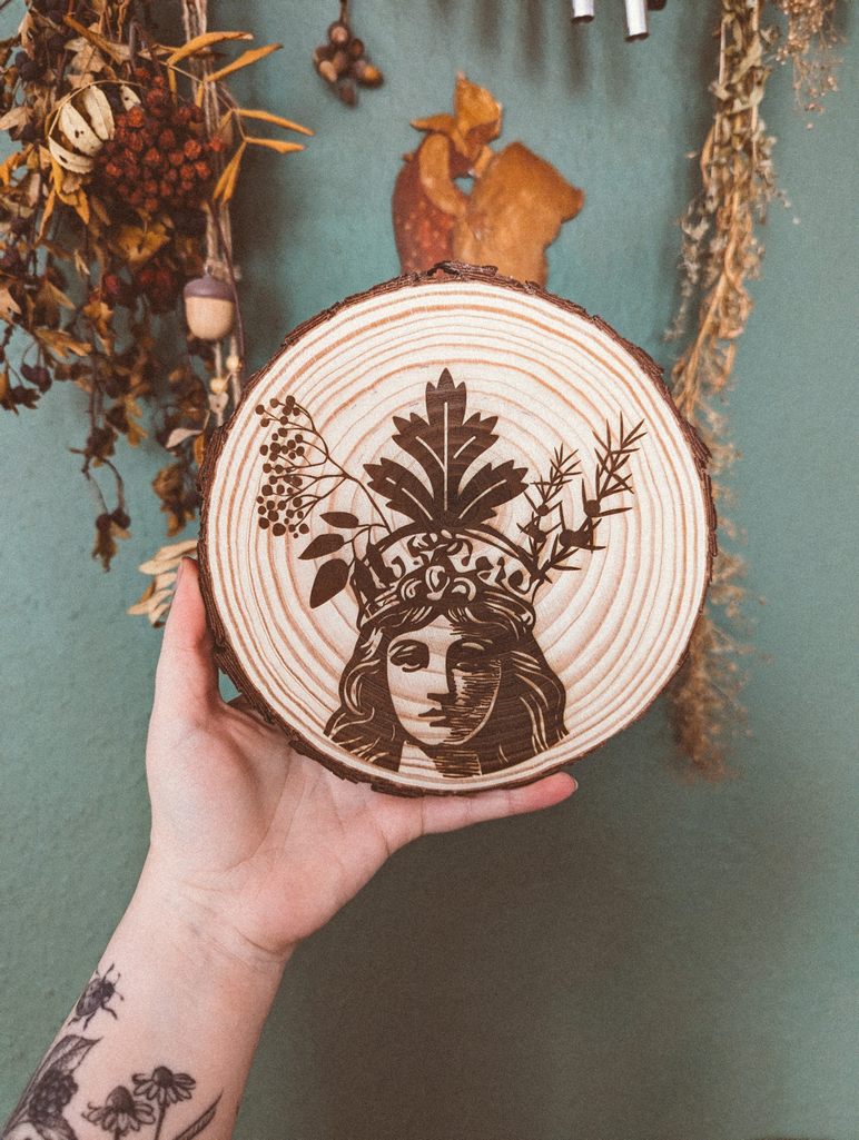 Eine Hand hält eine Baumscheibe: Eine Baumscheibe in der das Portait einer Frau eingebrannt ist. Eine ihrer Gesichtshälften ist dunkler gezeichnet als die andere. Auf dem Kopf trägt sie eine Krone aus Holunder, Beifuß und Wacholder.