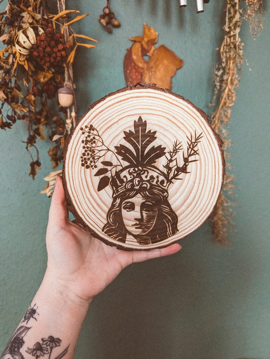 Eine Hand hält eine Baumscheibe: Eine Baumscheibe in der das Portait einer Frau eingebrannt ist. Eine ihrer Gesichtshälften ist dunkler gezeichnet als die andere. Auf dem Kopf trägt sie eine Krone aus Holunder, Beifuß und Wacholder.