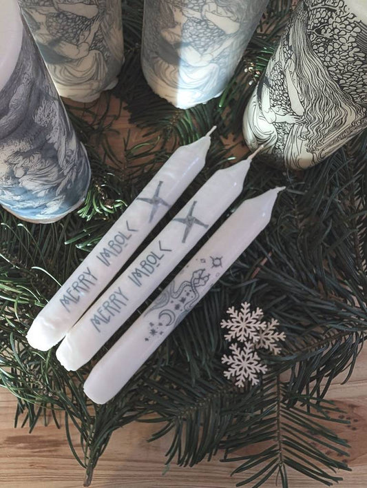 Mehrere weiße Kerzen mit verschiedenen Motiven stehen oder liegen auf Tannenzweigen.