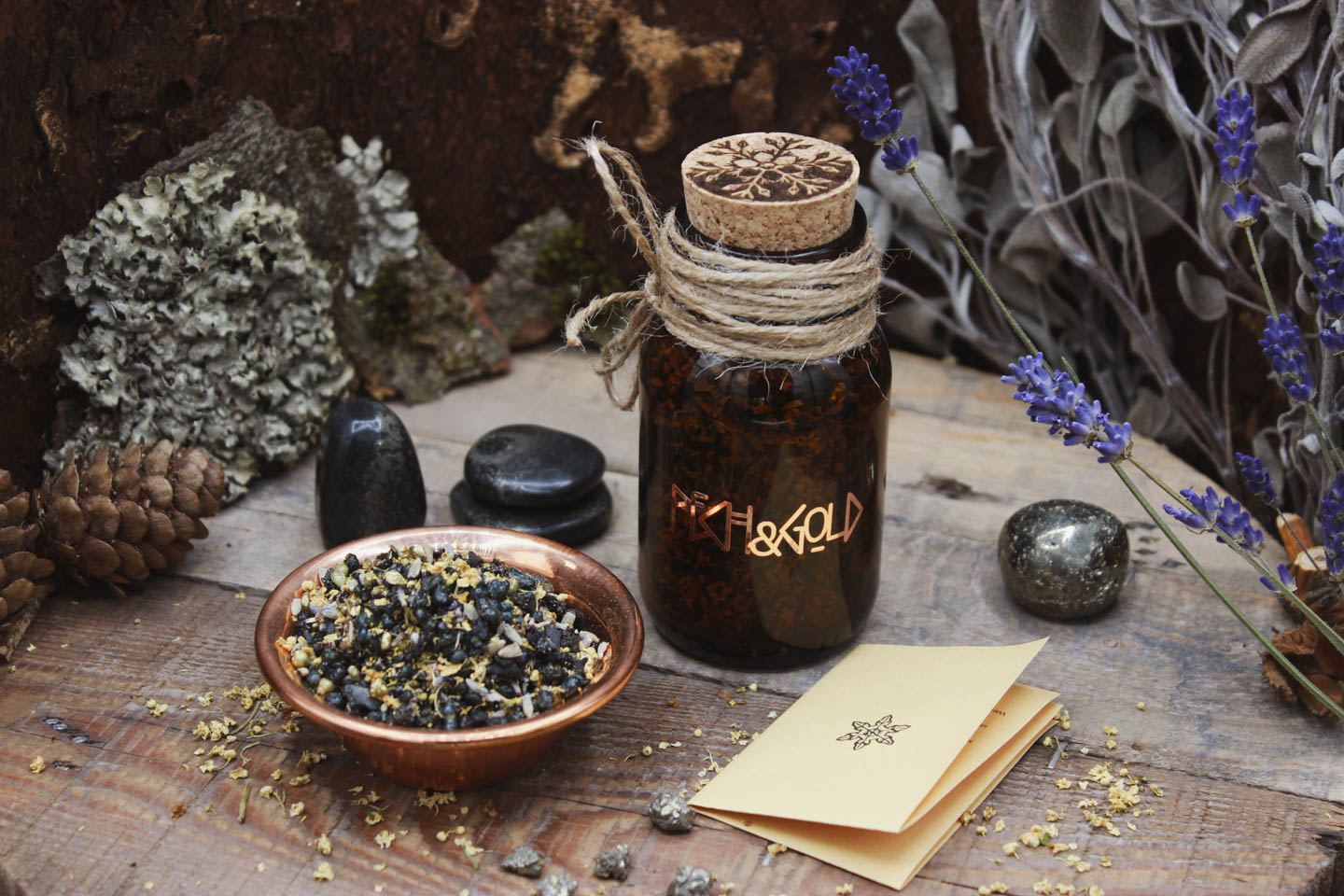 Räucherwerk in einem Braunglas mit Korken. Darauf steht in kupferner Schrift Pech & Gold. Daneben steht eine Kupferschale gefüllt mit Räucherwerk. Drumherum sind verschiedene Steine verteilt. Pyrit in Schiefer und kleine Pyrit-Stücke. Sowie einzelne kleine Holunderblüten. Ein kleiner Informationszettel liegt zusammengefaltet neben dem Glas.