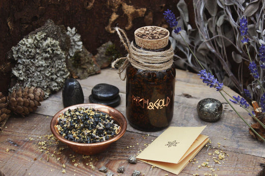 Räucherwerk in einem Braunglas mit Korken. Darauf steht in kupferner Schrift Pech & Gold. Daneben steht eine Kupferschale gefüllt mit Räucherwerk. Drumherum sind verschiedene Steine verteilt. Pyrit in Schiefer und kleine Pyrit-Stücke. Sowie einzelne kleine Holunderblüten. Ein kleiner Informationszettel liegt zusammengefaltet neben dem Glas.