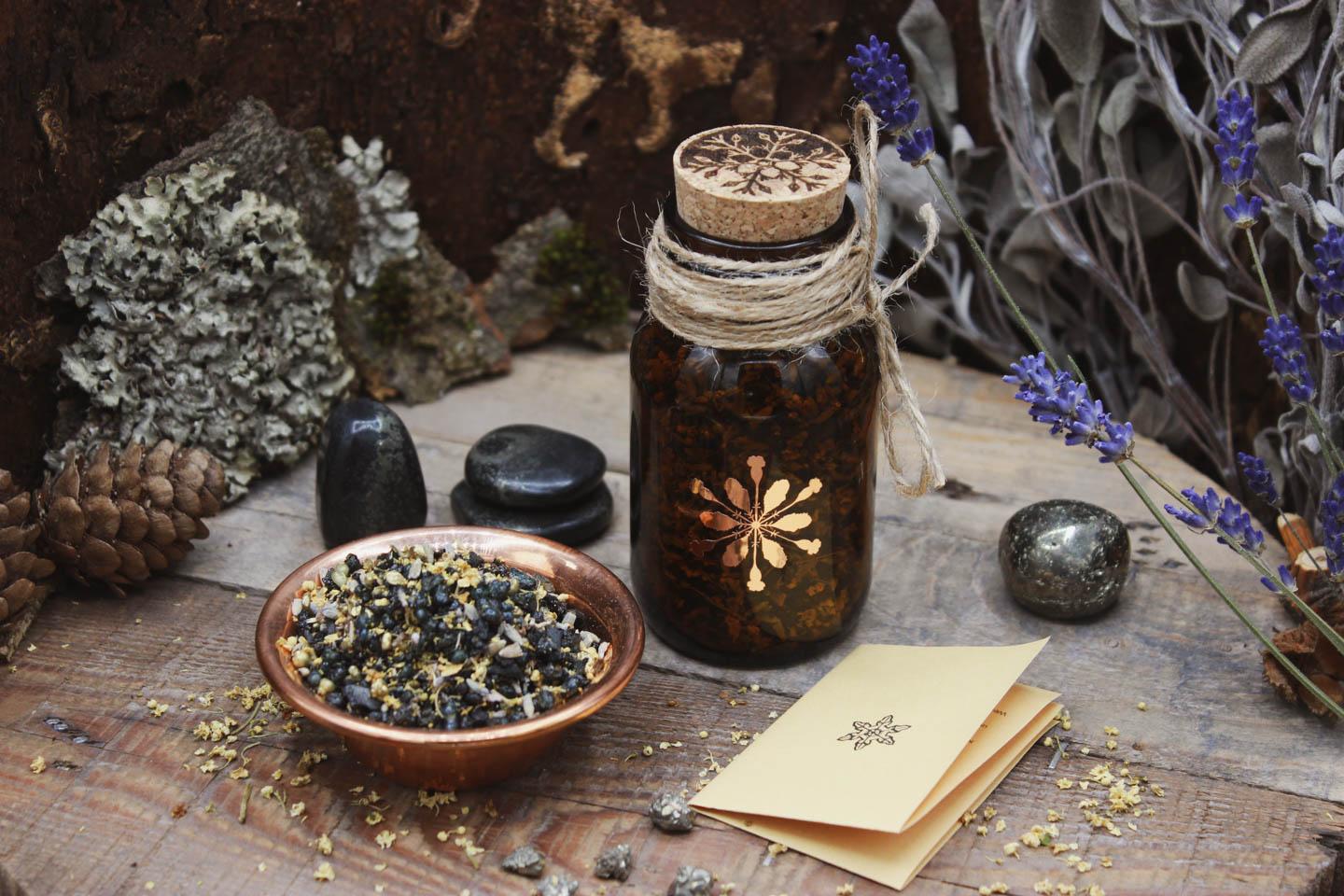 Räucherwerk in einem Braunglas mit Korken. Darauf ist das kupferne Hollenkraut-Logo zu sehen. Daneben steht eine Kupferschale gefüllt mit Räucherwerk. Drumherum sind verschiedene Steine verteilt. Pyrit in Schiefer und kleine Pyrit-Stücke. Sowie einzelne kleine Holunderblüten. Ein kleiner Informationszettel liegt zusammengefaltet neben dem Glas.