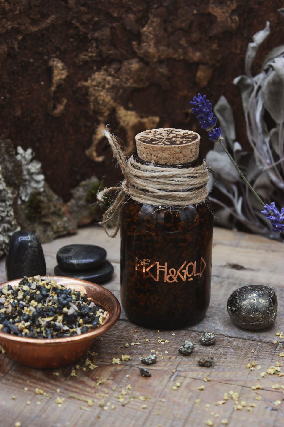 Räucherwerk in einem Braunglas mit Korken. Darauf steht in kupferner Schrift Pech & Gold. Daneben steht eine Kupferschale gefüllt mit Räucherwerk. Drumherum sind verschiedene Steine verteilt. Pyrit in Schiefer und kleine Pyrit-Stücke. Sowie einzelne kleine Holunderblüten.